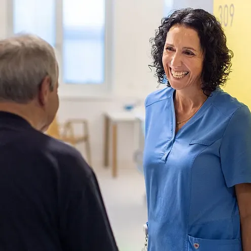 Pflegekraft in blauer Uniform lächelt einem älteren Patienten zu in einem hellen Krankenhausflur, gelbes Schild im Hintergrund.