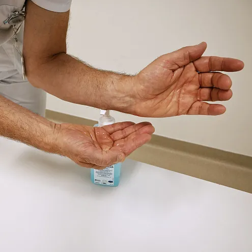 Eine Person zeigt ihre offenen Hände über einem weißem Tisch, neben einer großen Flasche mit Desinfektionsmittel. Der Fokus liegt auf der Desinfektion der Hände.