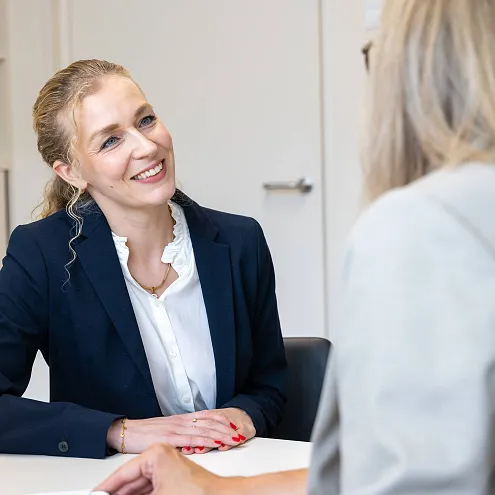 Two women in business attire having a professional discussion in an office setting. One woman is smiling and the other has her back to the camera.
