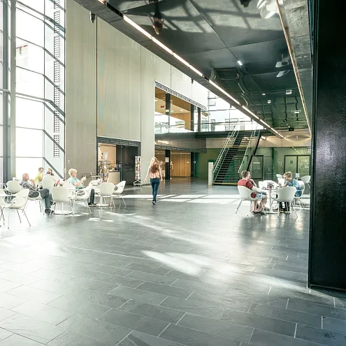 Modernes Foyer mit großen Fenstern, durch die viel Tageslicht einfällt. Menschen sitzen an Tischen, während andere durch den Raum gehen. Helle, offene Atmosphäre.