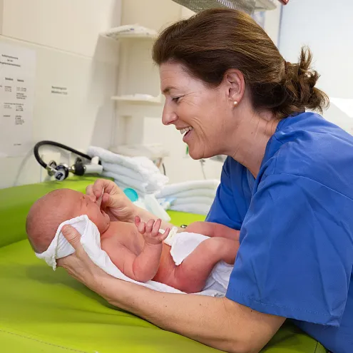 Krankenhausumgebung mit einer Krankenschwester, die ein neugeborenes Baby fürsorglich auf einem grünen Tisch hält. Beide wirken zufrieden und entspannt.