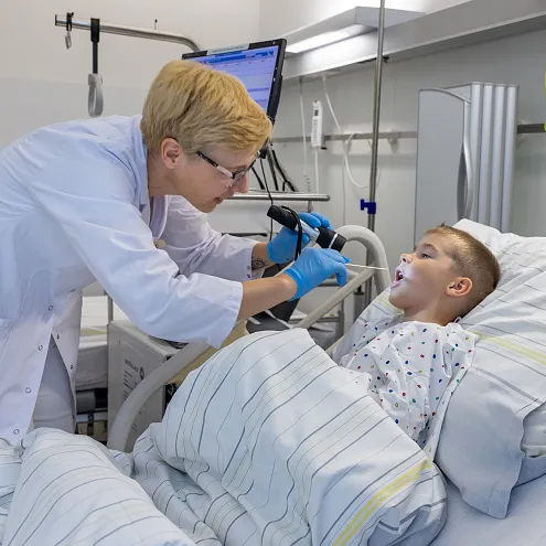A doctor examines a child in a hospital bed, using a light to look inside the child's mouth. The child wears a hospital gown. Medical equipment surrounds them.