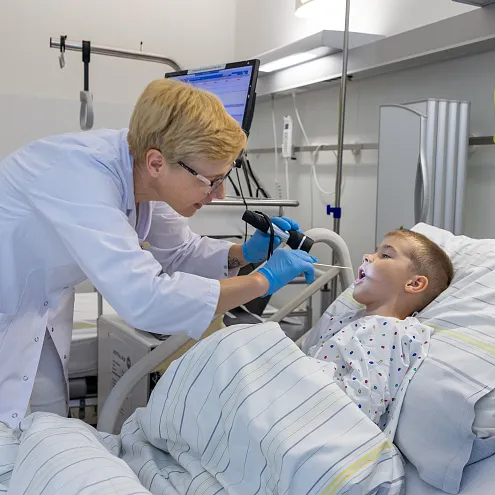 Eine Ärztin untersucht mit einem speziellen Gerät den Hals eines Jungen, der im Krankenhausbett liegt. Der Junge trägt ein Krankenhaushemd und wirkt ruhig.