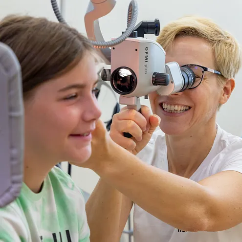 Eine Frau untersucht mit einem speziellen Gerät die Ohren eines jungen Mädchens, das lächelt. Beide befinden sich in einer medizinischen Untersuchungsumgebung.
