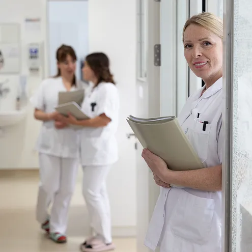 Drei Personen in weißer Arbeitskleidung stehen in einem hellen Raum. Eine Person im Vordergrund hält Akten und lächelt in die Kamera. Zwei stehen im Hintergrund.