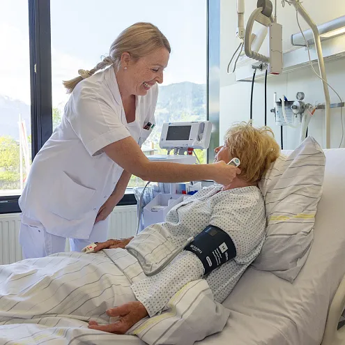 Eine Krankenschwester kümmert sich um eine Patientin in einem Krankenhauszimmer. Die Patientin liegt im Bett, während die Schwester lächelt und freundlich hilft.