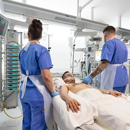 Zwei medizinische Fachkräfte in blauen Kitteln betreuen einen Patienten auf einer Intensivstation. Der Raum ist mit moderner medizinischer Ausrüstung ausgestattet.