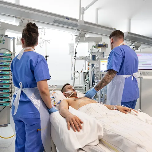 Zwei medizinische Fachkräfte in blauen Kitteln betreuen einen Patienten auf einer Intensivstation. Der Raum ist mit moderner medizinischer Ausrüstung ausgestattet.