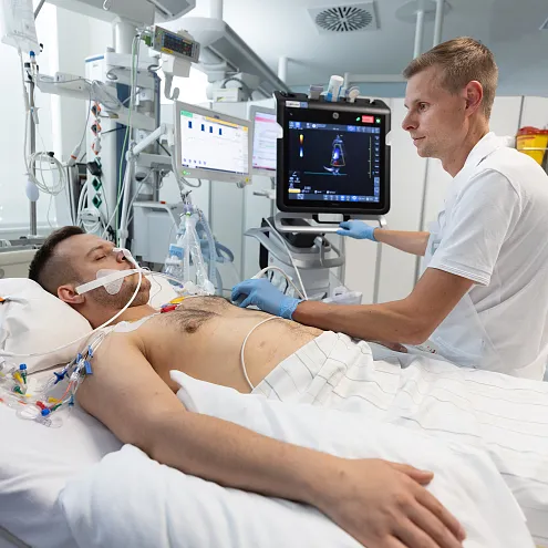 Ein:e Ärzt:in überwacht einen liegenden Patient:in im Krankenhausbett mit angeschlossenen Monitoren und medizinischen Geräten in einem hellen Krankenzimmer.