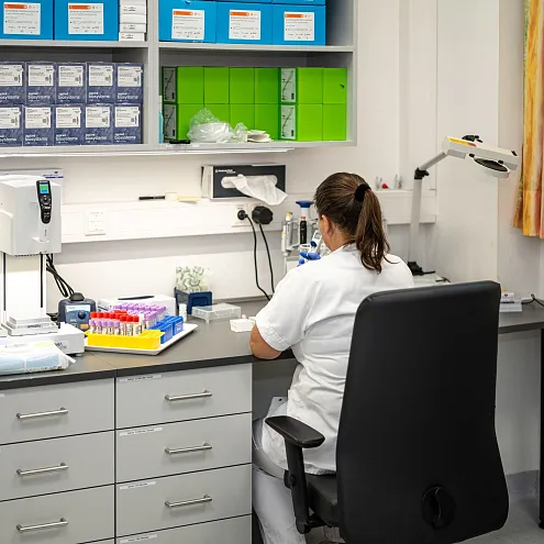 Eine Person in weißem Kittel arbeitet in einem Labor, umgeben von Regalen mit Boxen und Labormaterialien. Sie sitzt an einem Tisch mit Reagenzgläsern.