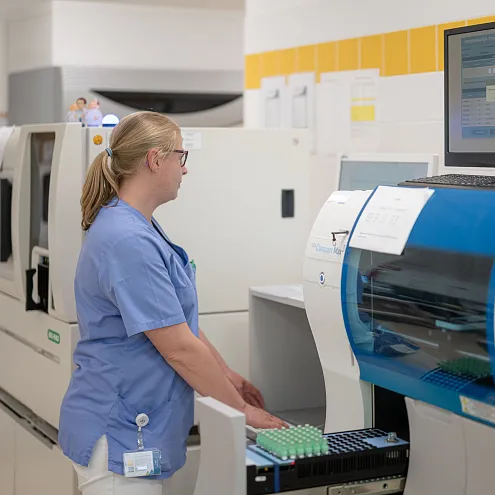 Eine Frau in blauer Arbeitskleidung steht in einem Labor vor einem medizinischen Analysegerät und bedient einen Computer. Regale sind im Hintergrund zu sehen.
