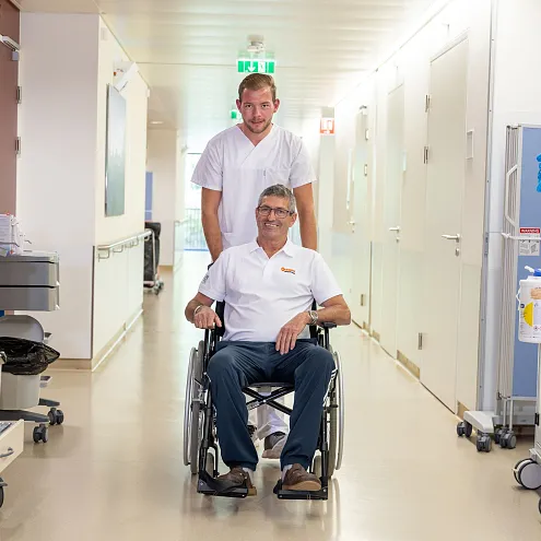 Ein Krankenpfleger schiebt einen lächelnden Mann im Rollstuhl durch einen hellen Krankenhausflur. Beide tragen weiße Kleidung, umgeben von medizinischem Equipment.