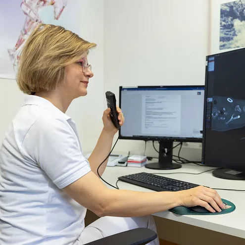 Eine medizinische Fachkraft in weißem Kittel betrachtet CT-Bilder auf zwei Monitoren und nutzt ein Handmikrofon zur Dokumentation oder Kommunikation.