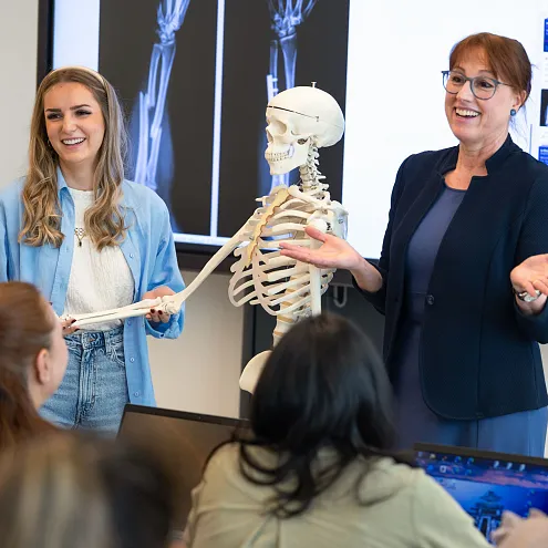 Zwei Frauen präsentieren vor Studierenden in einem Klassenraum. Ein Skelett und Röntgenbilder sind im Hintergrund zu sehen. Alle tragen lässige Kleidung.