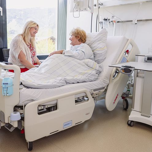 Eine Frau sitzt lächelnd auf einem Stuhl neben einem Krankenhausbett, in dem eine andere Frau liegt. Durch ein großes Fenster scheint Tageslicht herein.