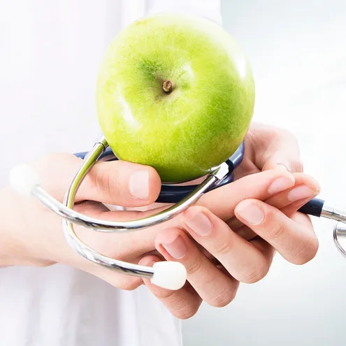 Eine Person hält einen grünen Apfel und ein Stethoskop in den Händen. Der Hintergrund ist unscharf und zeigt ein helles, modernes Umfeld.