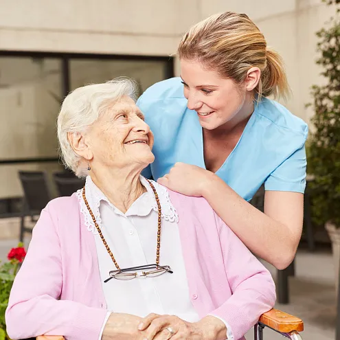 Eine lächelnde ältere Frau im Rollstuhl wird von einer freundlichen jungen Pflegekraft betreut. Im Hintergrund sind Pflanzen und ein Gebäude zu sehen.