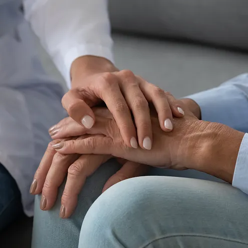 Gehobene Perspektive auf zwei Menschen, die miteinander sprechen. Eine Person hält beruhigend die Hand der anderen, während sie Notizen macht. Vertrauliche Atmosphäre.