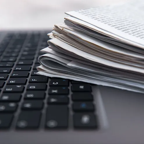 Ein aufgeschlagener Laptop ist zu sehen, auf dessen Tastatur ein Stapel Zeitungen liegt. Die Szenerie deutet auf ein modernes Arbeitsumfeld hin.