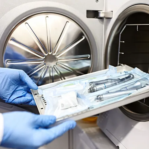 Ein Mensch legt ein Tablett mit sterilen medizinischen Instrumenten in einen Autoklav zur Sterilisation. Die Person trägt blaue Schutzkleidung und -handschuhe.