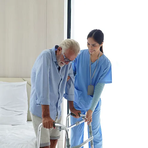 Eine Krankenschwester unterstützt einen älteren Mann mit einem Gehhilfe beim Gehen in einem hellen Raum. Beide wirken konzentriert und vertraut miteinander.