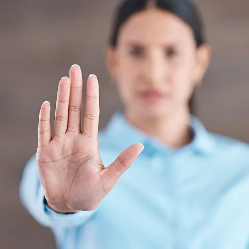 Eine Person im unscharfen Hintergrund hält die Hand mit ausgestreckter Handfläche nach vorne, als Zeichen von Stopp oder Ablehnung, vor einem neutralen Hintergrund.