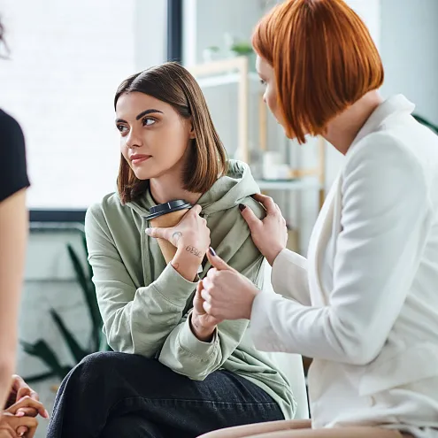 Drei Frauen in einem hellen Raum führen ein ernstes Gespräch. Eine Frau in Grün hält ihre Hände verschränkt, während die anderen beiden aufmerksam zuhören. 