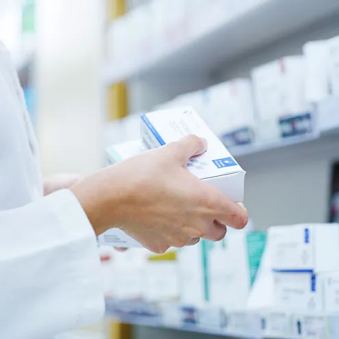 Ein Apotheker in weißem Kittel hält eine Medikamentenverpackung in der Hand, stehend vor einem Regal mit weiteren Arzneimitteln in einer Apotheke.