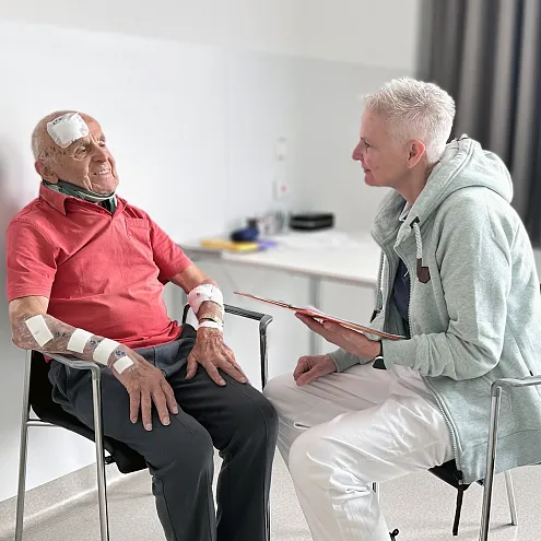 Ein älterer Mann mit Verbänden sitzt in einem Stuhl und wird von einem Mann in Arzthelferuniform mit einem Tablet in der Hand freundlich angesprochen.