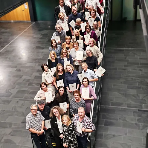 Eine große Gruppe von Menschen steht auf einer Treppe in einem Gebäude. Sie halten Zertifikate und lächeln in die Kamera. Es herrscht eine fröhliche Atmosphäre.