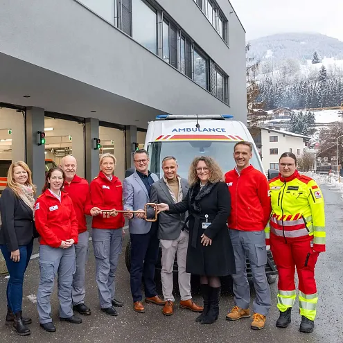 Eine Gruppe von Menschen steht vor einem modernen Rettungsgebäude. Im Hintergrund sind ein Rettungsfahrzeug und schneebedeckte Bäume zu sehen.
