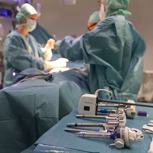 Chirurgische Instrumente liegen auf einem Tisch im Vordergrund, während im Hintergrund Ärzte in OP-Kleidung eine Operation durchführen, umgeben von medizinischen Geräten.