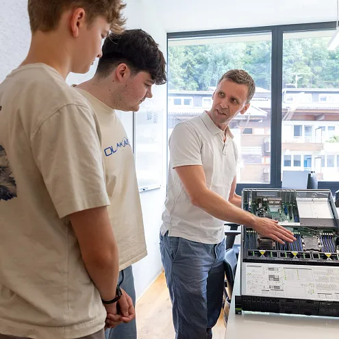 Ein Mann erklärt zwei jungen Männern vor einem Fenster mit Bergblick die technischen Details eines elektronischen Geräts auf einem Tisch.