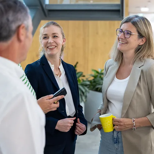 Drei Personen in Geschäftskleidung unterhalten sich fröhlich in einem modernen Bürogebäude. Eine Frau hält einen gelben Becher. Eine Treppe ist im Hintergrund.