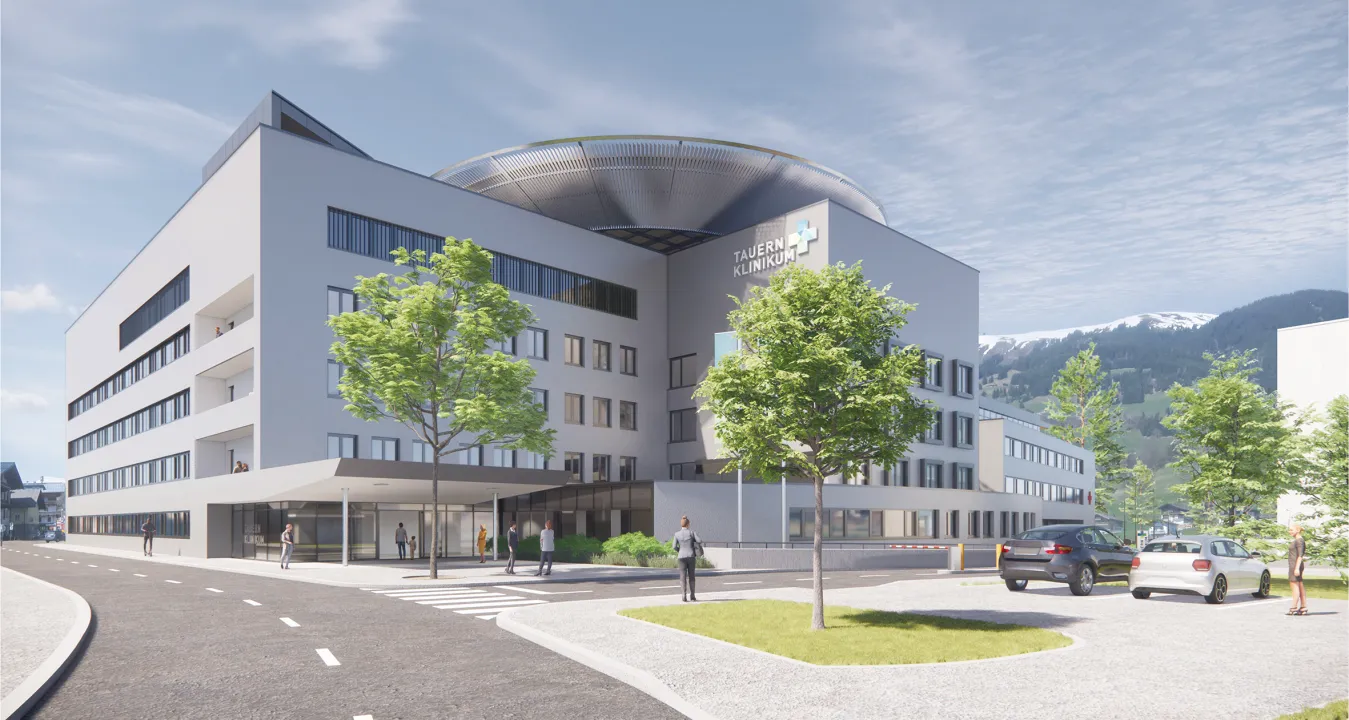 Moderne Klinik mit Glasfassade und großem Eingangsbereich, umgeben von Bäumen und Parkplätzen. Im Hintergrund sind bewaldete Hügel und blauer Himmel sichtbar.