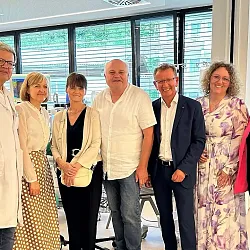 Eine Gruppe von Menschen steht lächelnd in einem modernen, gut beleuchteten Raum. Einige tragen formelle Kleidung, andere lässige Kleidung. Der Raum hat große Fenster.