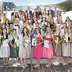 Eine große Gruppe lächelnder Menschen in traditioneller Kleidung steht auf Treppen im Freien, einige halten Sonnenblumen und Dokumente in den Händen.