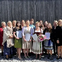 Eine Gruppe von Menschen posiert in traditioneller Tracht lächelnd vor einer Holzwand. Einige halten Urkunden und Blumensträuße in den Händen.