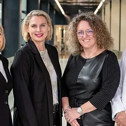 Vier Personen in formeller Kleidung, drei Frauen und ein Mann in Arztkittel, stehen in einem modernen Korridor und lächeln in die Kamera.