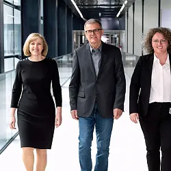 Drei Personen in formeller Kleidung gehen lächelnd einen modernen Flur entlang. Im Hintergrund sind große Fenster mit Blick auf eine Landschaft zu sehen.