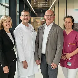 Vier Männer und zwei Frauen stehen in einem modernen Gebäude. Sie tragen teils Kittel und teils Zivilkleidung und lächeln in die Kamera.