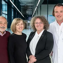 Vier lächelnde Personen, drei in Freizeitkleidung, eine im weißen Kittel, stehen in einem Gebäudekorridor mit Glaswänden und moderner Beleuchtung.