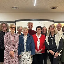Gruppe von Menschen steht lächelnd in einem Raum vor einem Whiteboard. Die Personen tragen unterschiedliche Kleidung, von formell bis casual.