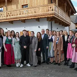 Eine Gruppe von Menschen in traditioneller und formeller Kleidung posiert vor einem alpinen Gebäude mit Holzbalkon und blauer Himmelskulisse.