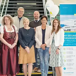 Eine Gruppe von sieben Personen steht vor einem Werbeplakat des Tauern Klinikums. Sie lächeln und wirken professionell gekleidet. Luftballons sind sichtbar.