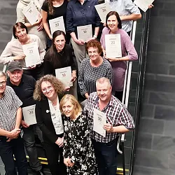 Eine Gruppe von Menschen steht auf einer Treppe, sie halten Zertifikate und lächeln in die Kamera. Sie tragen verschiedene Freizeitkleidung.