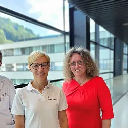 Drei Personen stehen in einem modernen Gebäude mit großen Fenstern. Zwei tragen weiße Arbeitskleidung, die dritte Person trägt ein rotes Oberteil.