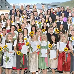 Gruppenfoto von Menschen in traditioneller Kleidung, die lächelnd Sonnenblumen und Urkunden halten. Die Kulisse zeigt Gebäude und Berge.