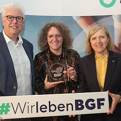 Drei Personen präsentieren stolz eine Auszeichnung und halten ein Schild mit der Aufschrift '#WirlebenBGF'. Sie stehen vor einem Hintergrund mit Logos.