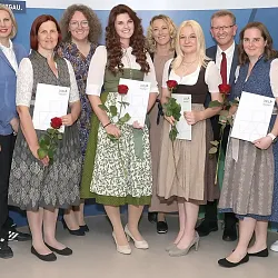 Eine Gruppe von 12 Personen in traditioneller bayerischer Tracht. Frauen halten Zertifikate und Rosen, sie lächeln in die Kamera. Hintergrund: blaues Banner.